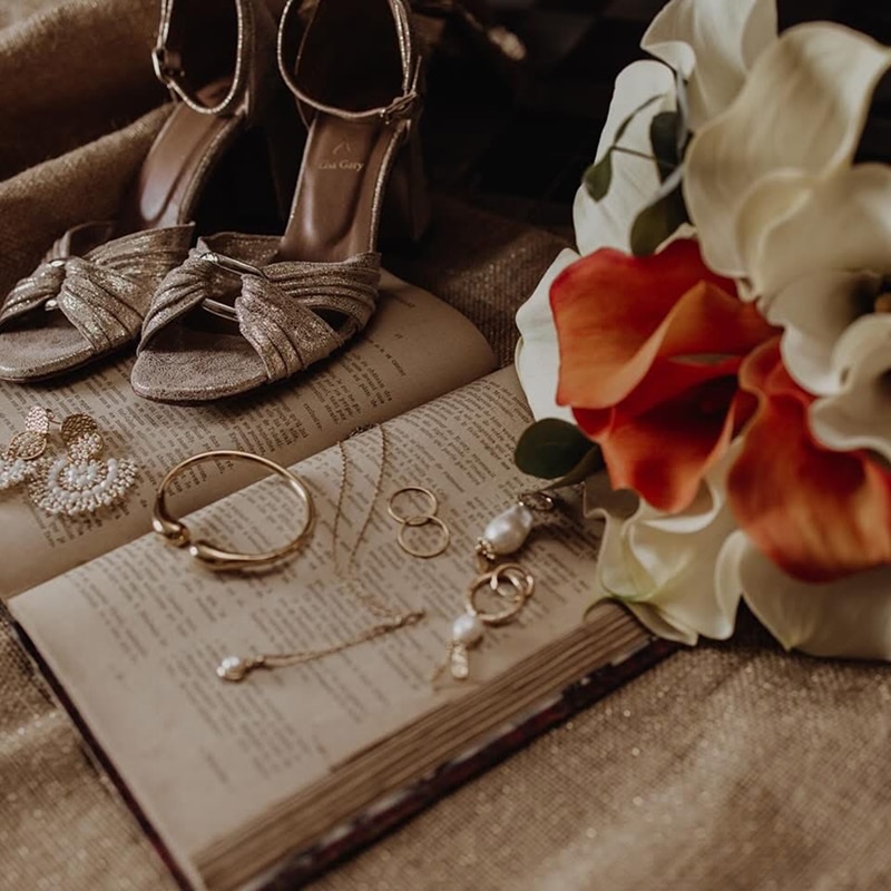 Chaussures et bijoux de mariée dorés sur un livre ancien Chaussures dorées "Lisa Gary", bijoux fins à perles et boucles d'oreilles sur un livre ouvert, près d'un bouquet de fleurs oranges et blanches.