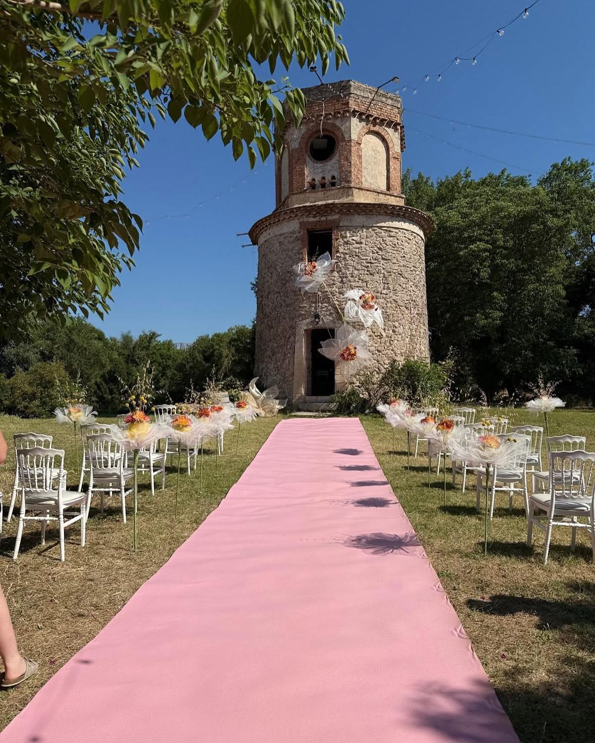 Allée de mariage avec tapis rose et compositions florales devant un moulin en pierre
