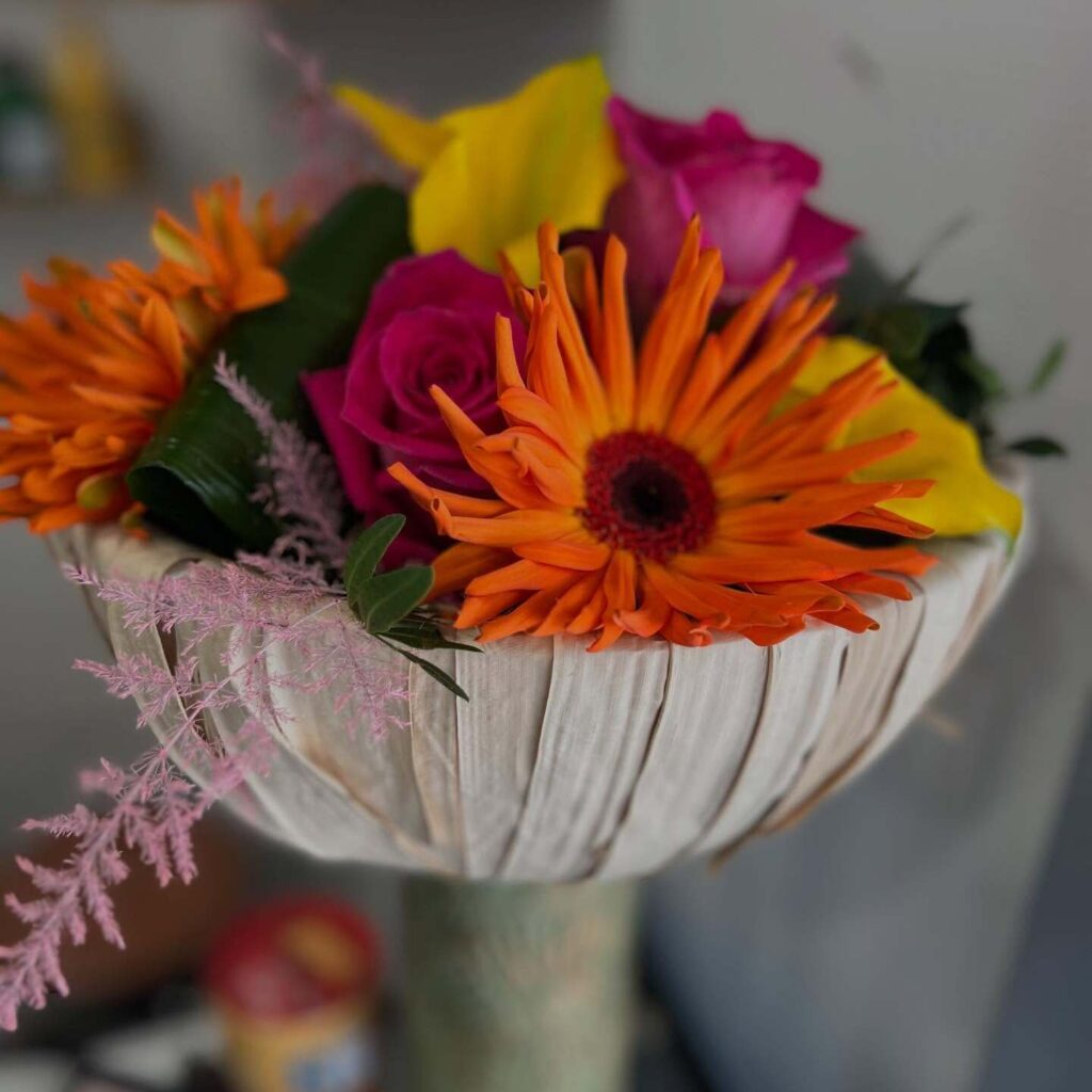Arrangement floral éclatant : Gerbera orange et Roses Vibrant bouquet de Gerbera orange, roses magenta, et calla jaune dans un contenant tressé de fibres naturelles, décoré de feuillage rose.