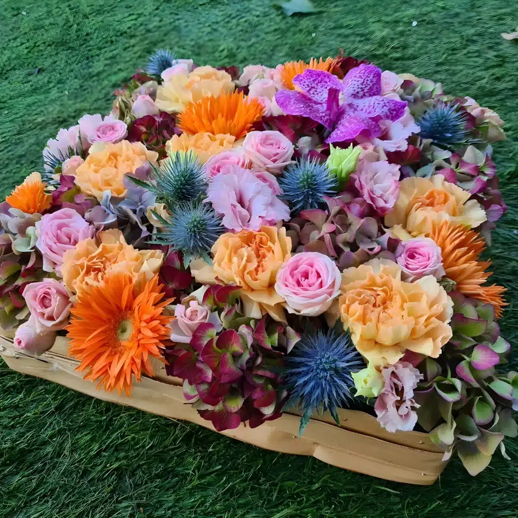 Fleurs tons chauds pour hommage : roses, orange et bleu intense. Composition florale cœur en tons chauds: roses pêche, gerbera orange, orchidée violette et chardons bleus sur gazon synthétique.